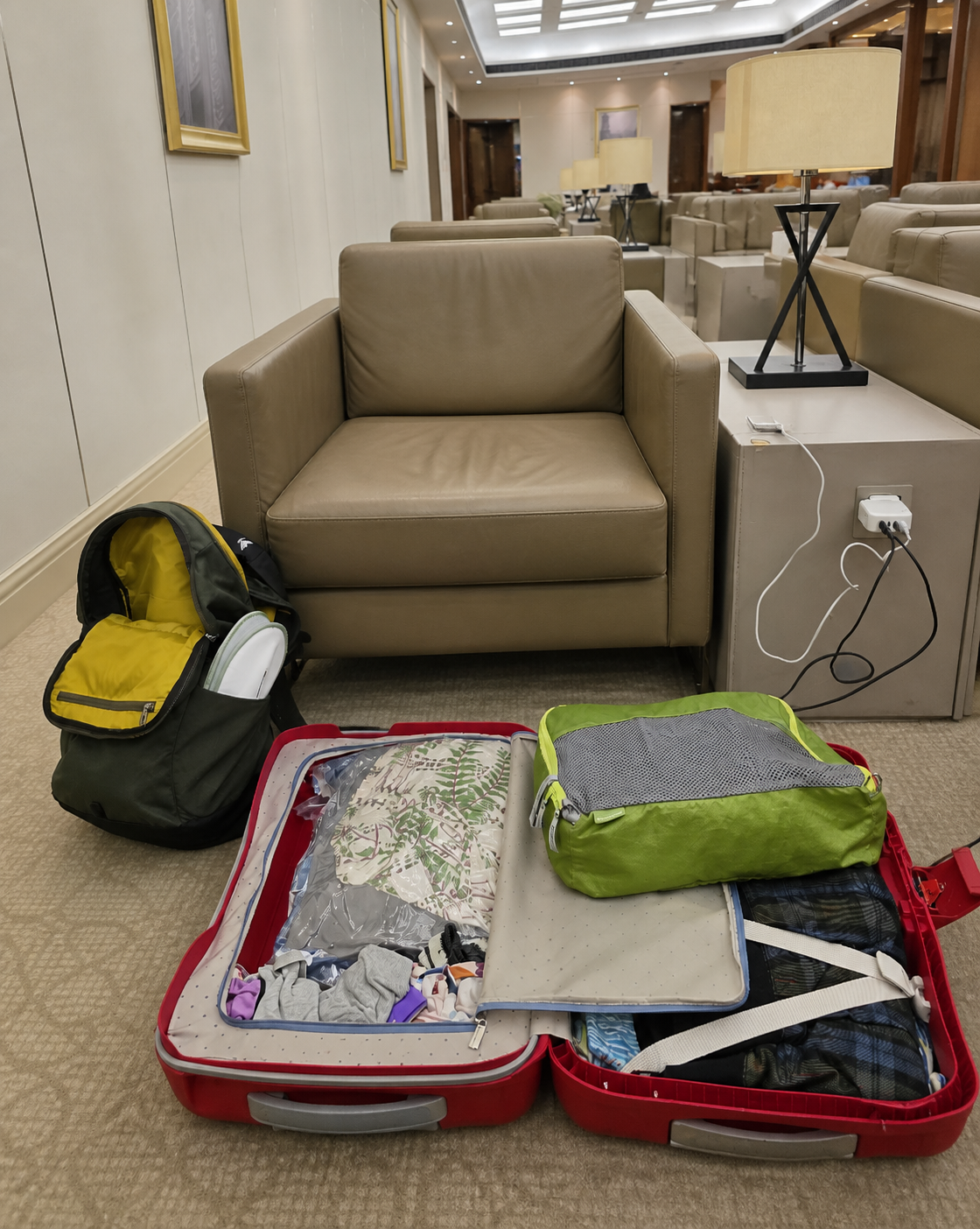 An open red suitcase, packing cube, carry-on bag and charging cable in an airport lounge
